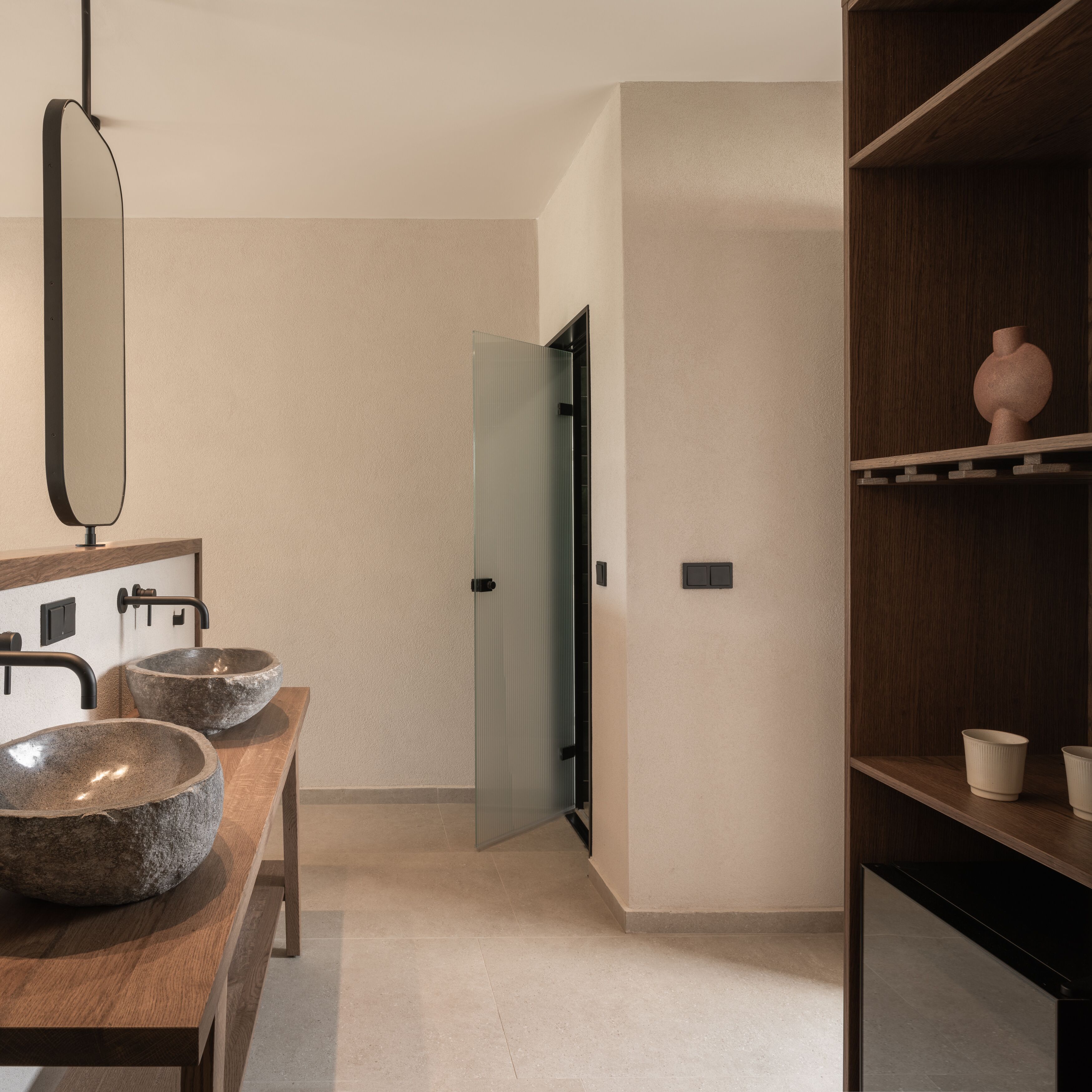 A modern bathroom with a wooden vanity, two stone sinks, a mirror, and a wooden shelf with decorative items.