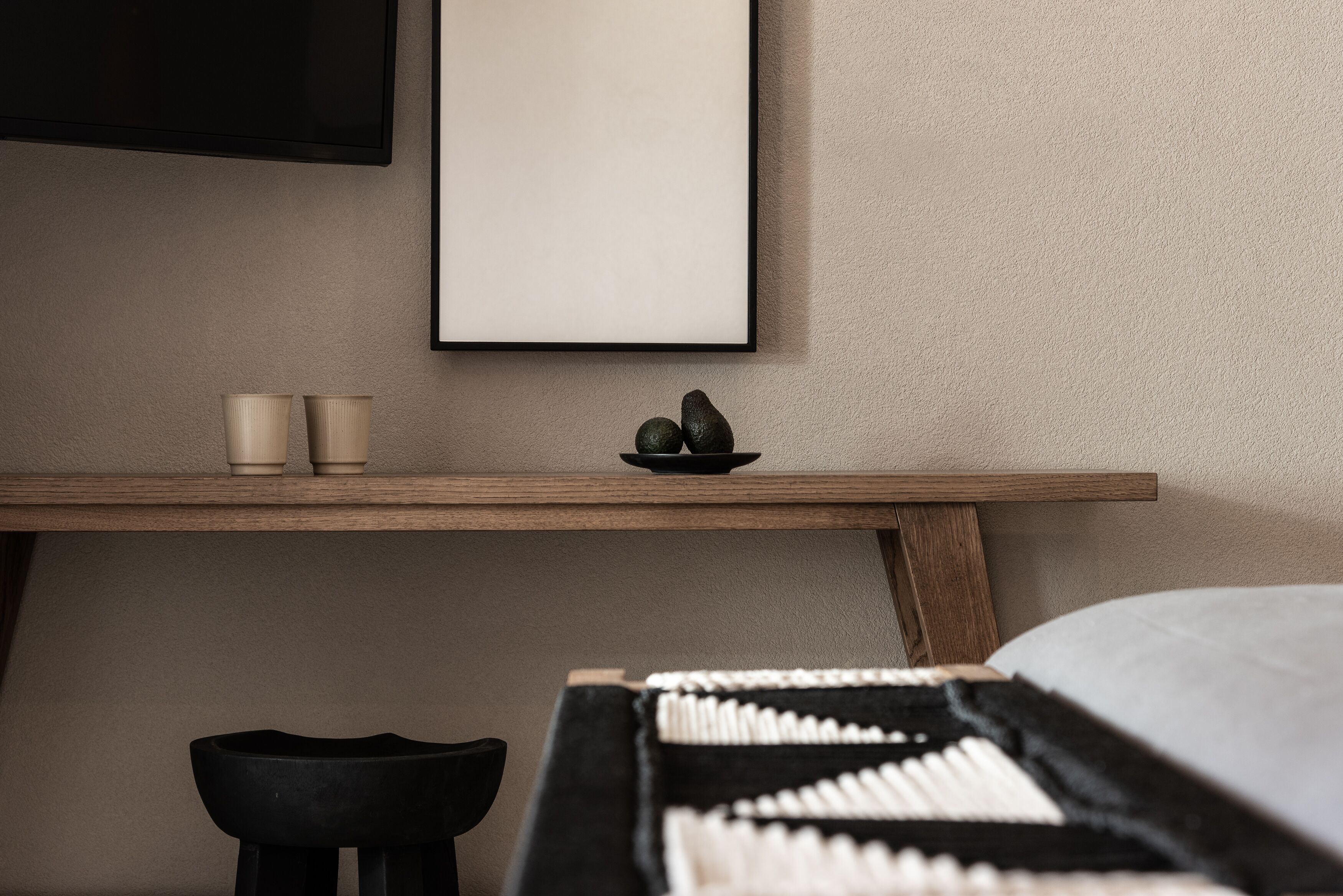A minimalist room with a bed, a wooden nightstand, a black stool, and a framed picture on the wall.
