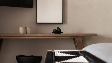 A minimalist room with a bed, a wooden nightstand, a black stool, and a framed picture on the wall.