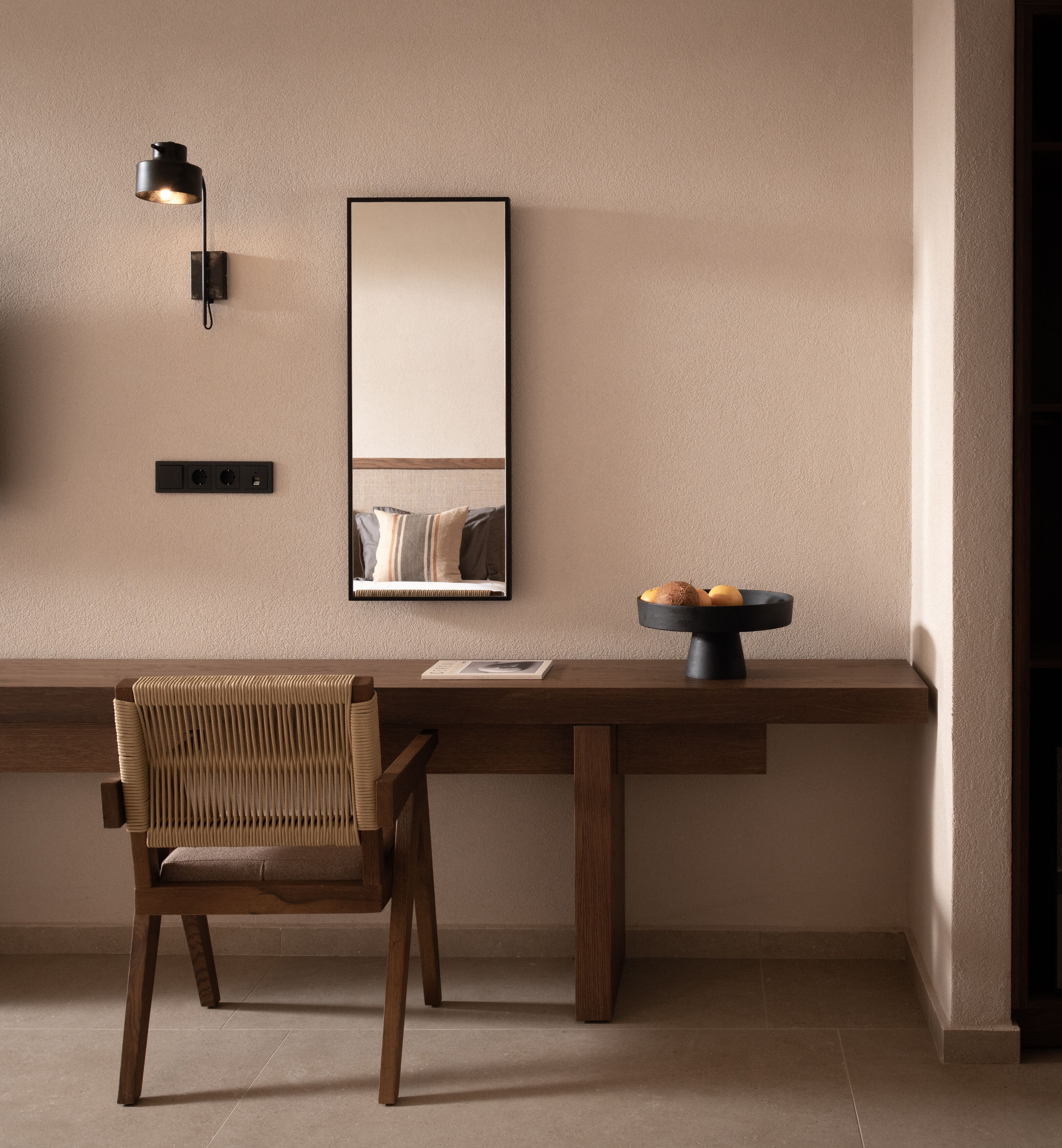 A modern interior with a wooden desk, a woven chair, a black-framed mirror, and a wall-mounted light fixture.