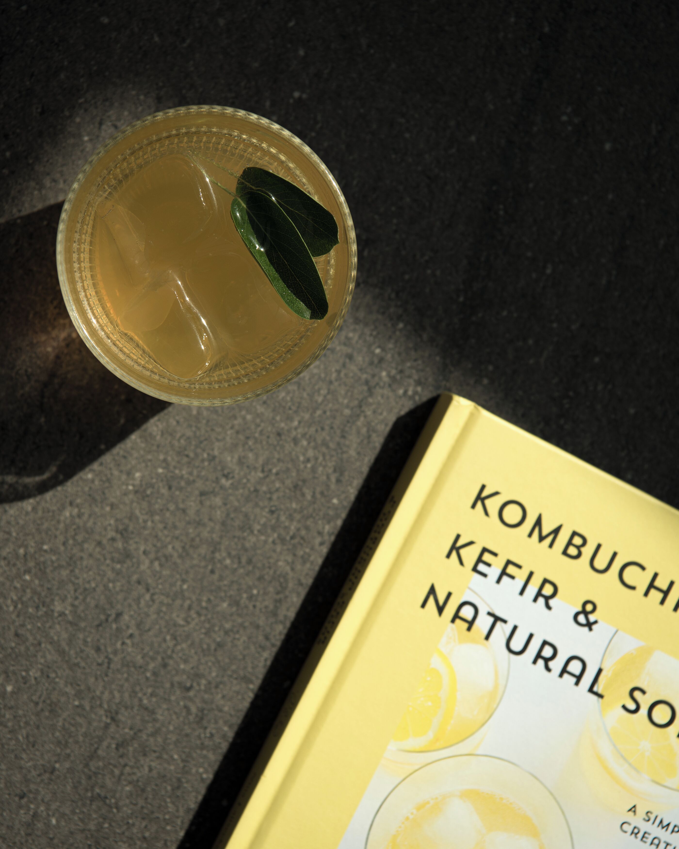 Un vaso de kombucha con una hoja verde, colocado junto a un libro titulado 'Kombuch Kefir & Natural Soda'.