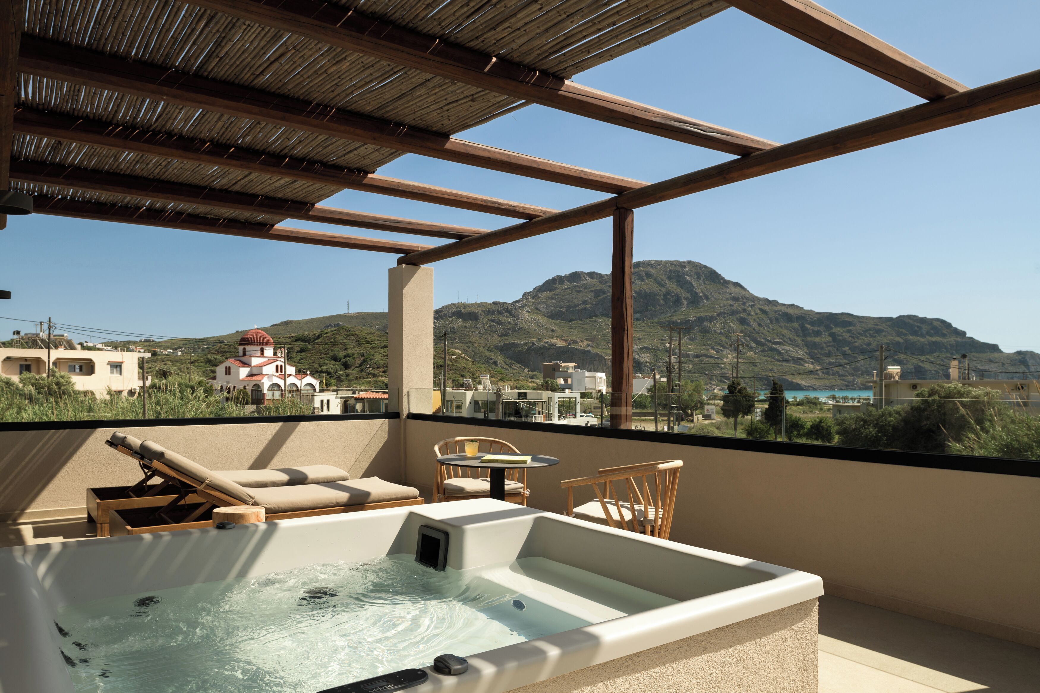 Terraza en la azotea con jacuzzi, sillones de descanso y una mesa, con vistas a un paisaje montañoso y edificios.