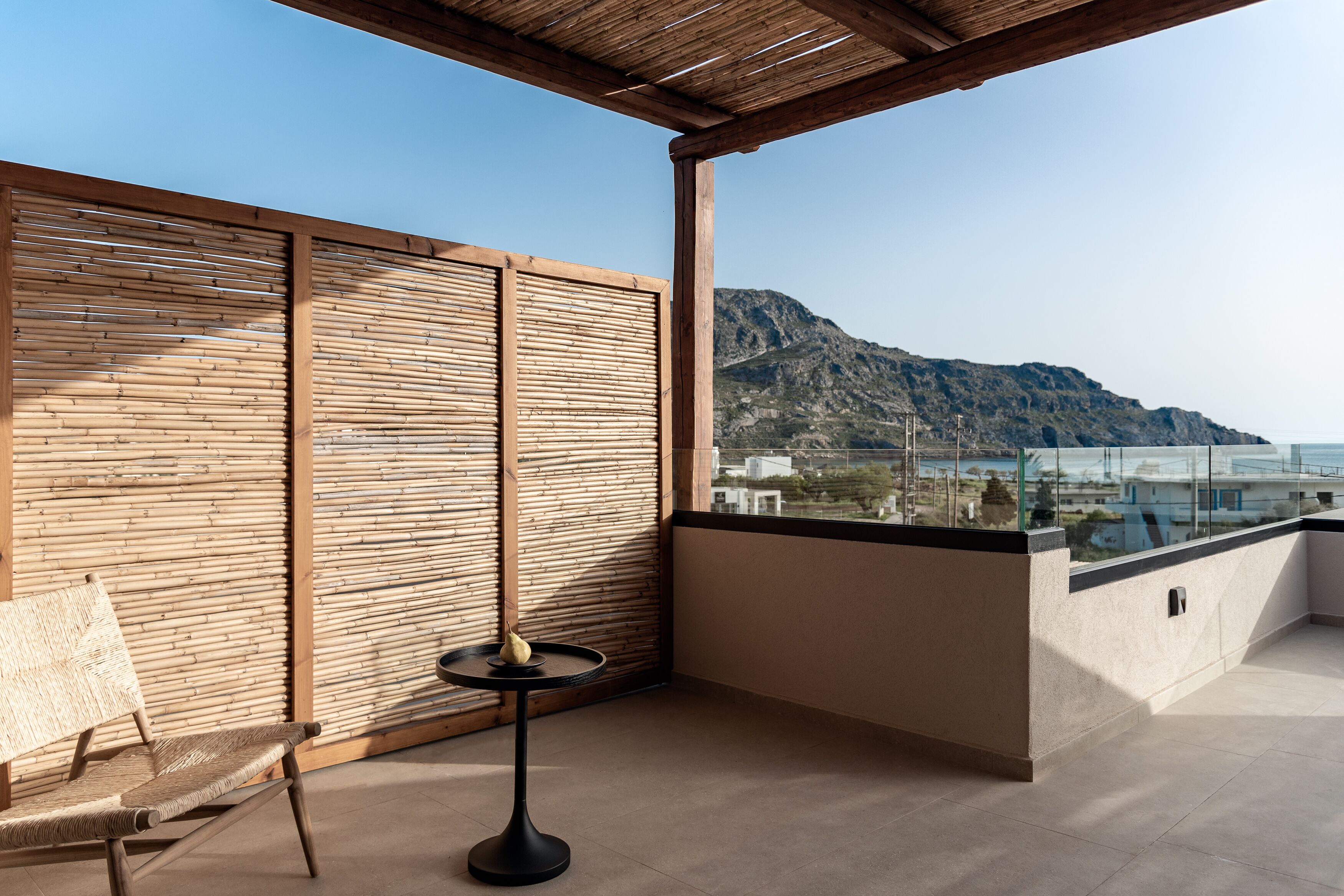 Un balcón con pared de madera, una silla de mimbre, una pequeña mesa redonda con una pera y vistas a montañas y edificios.