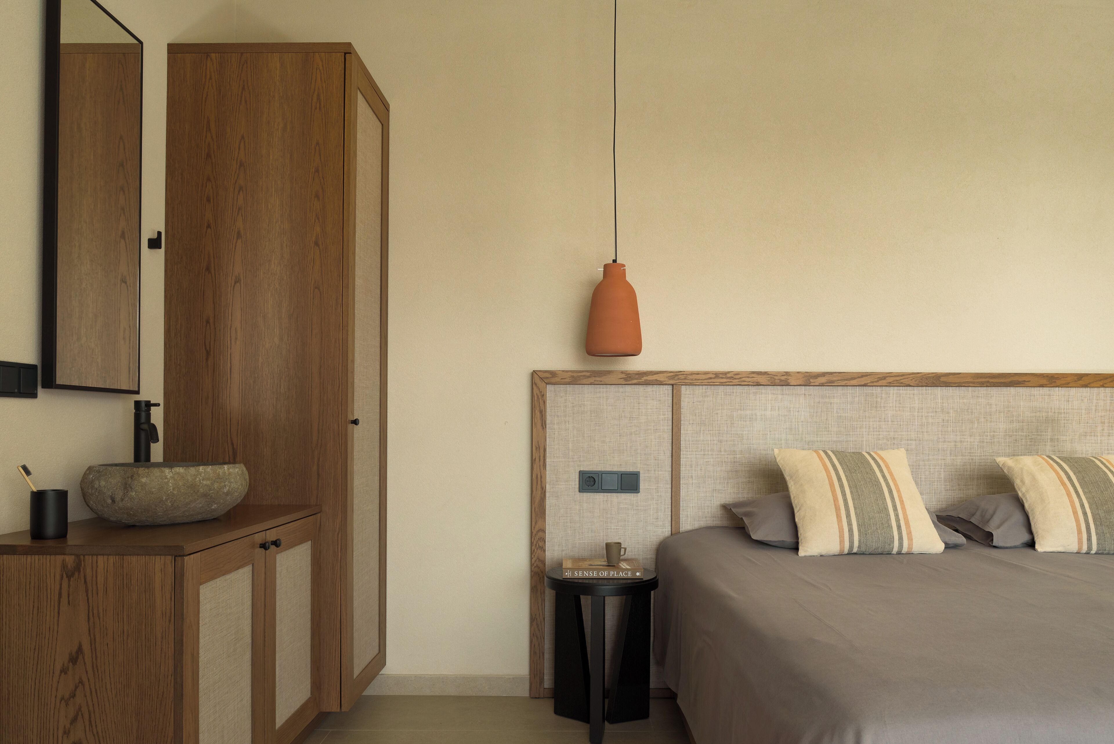 Ein Schlafzimmer mit einem Holzschrank, einem runden Waschbecken, einem kleinen Tisch mit Buch und Tasse sowie einem Bett mit gestreiften Kissen.