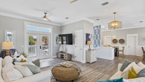 A spacious living room with a white sofa, a wicker ottoman, and a dining area with a kitchen in the background.