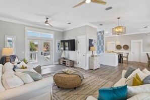 A spacious living room with a white sofa, a wicker ottoman, and a dining area with a kitchen in the background. - Panama City Beach House with Saltwater Pool and Spa | Directly Across from Beach (Panama City Beach)