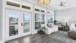 A spacious living room with a white sofa, a wicker ottoman, and a view of the ocean through large windows.