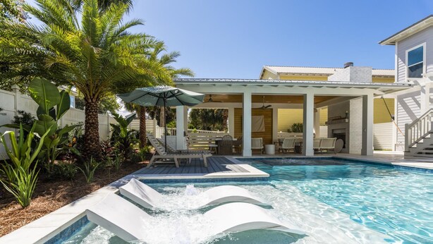 A swimming pool with a waterfall feature, surrounded by lounge chairs and umbrellas, with a covered patio area and a house in the background.