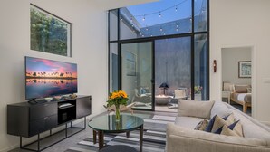 A modern living room with a flat-screen TV, a glass coffee table, a sofa, and a dining table with a vase of flowers. - Historic Post Office Loft w/firepit near Zoo (San Diego)