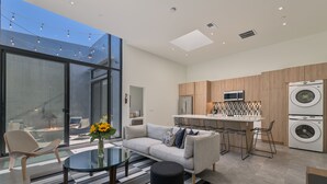 A modern kitchen with a microwave, refrigerator, and washing machine. There is a glass dining table with chairs and a sofa with cushions. - Historic Post Office Loft w/firepit near Zoo (San Diego)