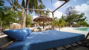 A bed with a blue mattress under a wooden canopy, a pool, and palm trees in the background. - The Mida Creek (Watamu)
