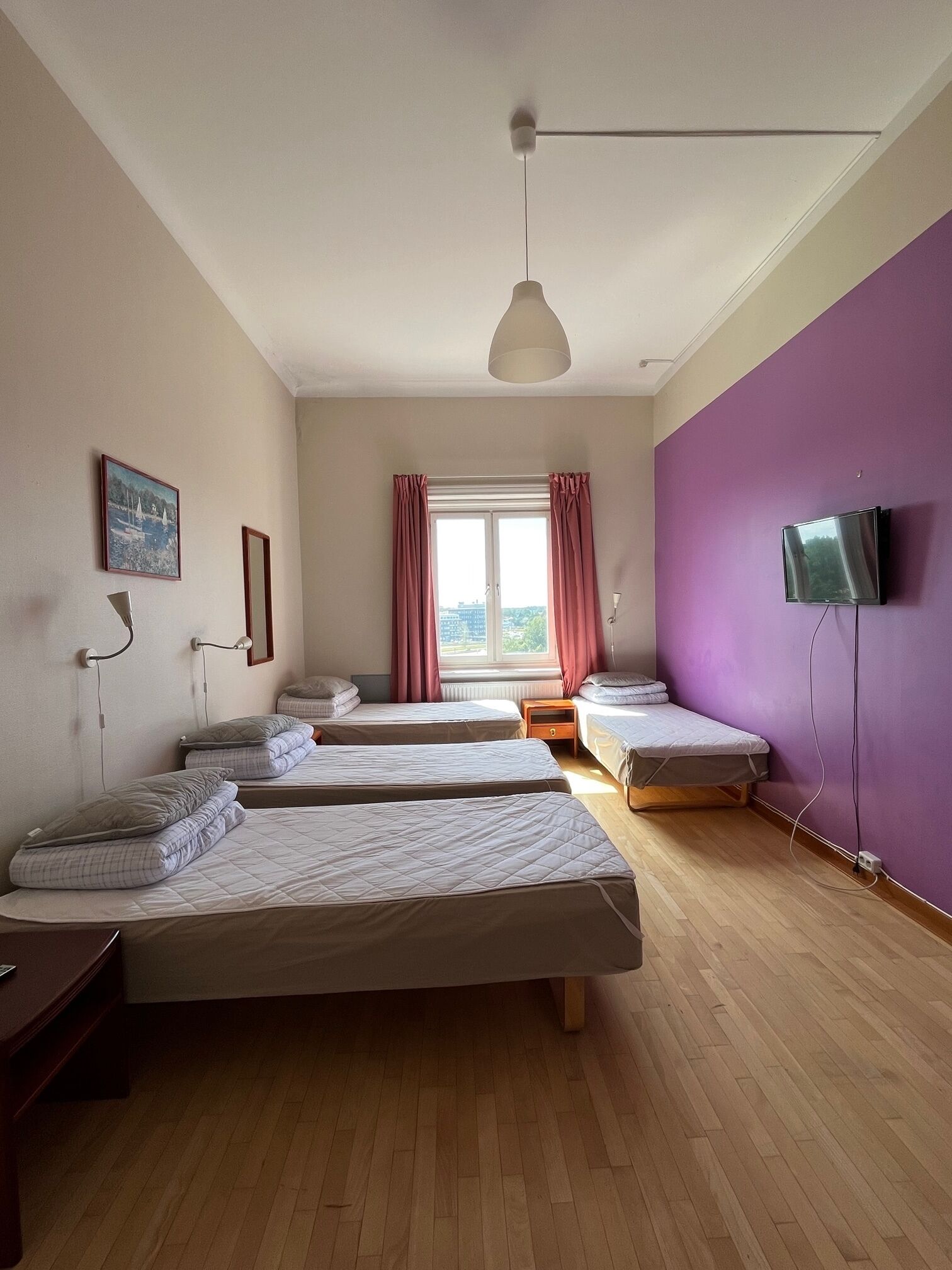 A room with four single beds, a ceiling fan, a window with curtains, a television mounted on the wall, and a framed picture on the wall.