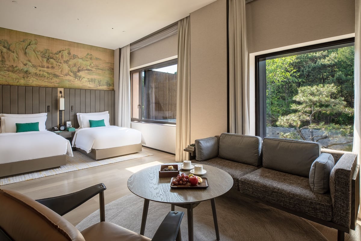Ett hotellrum med två sängar, en soffa, ett runt bord med frukt och ett stort fönster med utsikt över grönområden.