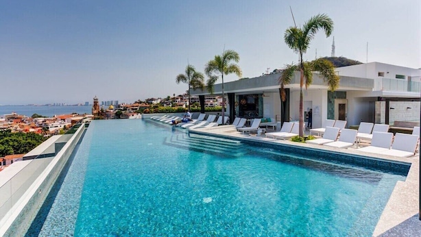 A swimming pool with lounge chairs and palm trees, overlooking a coastal cityscape.