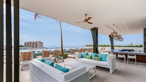 A modern outdoor lounge area with white furniture, a bar, and a view of the ocean and buildings.