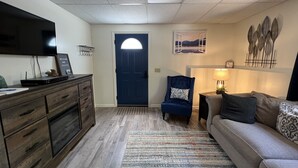 Un salon avec une porte bleu foncé, un téléviseur à écran plat posé sur un meuble en bois, un fauteuil bleu agrémenté d’un coussin à motifs, un canapé gris avec des coussins à motifs et un élément décoratif mural.