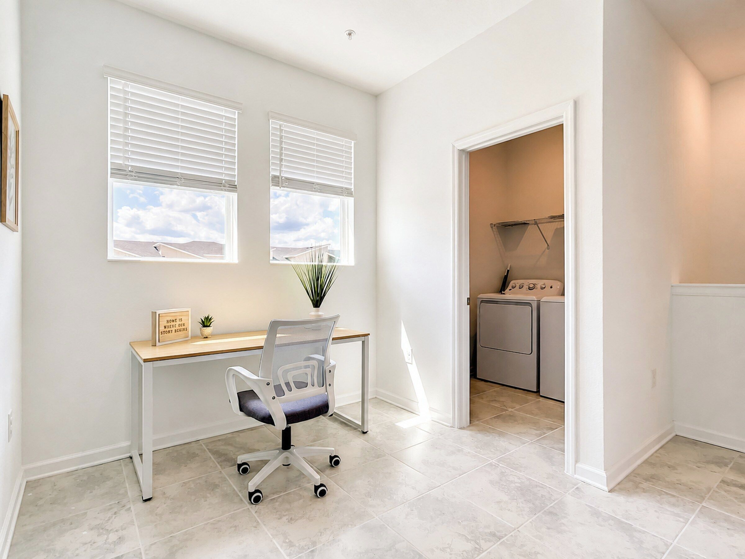 Una habitación con escritorio, silla, dos ventanas, una planta y zona de lavandería con lavadoras.
