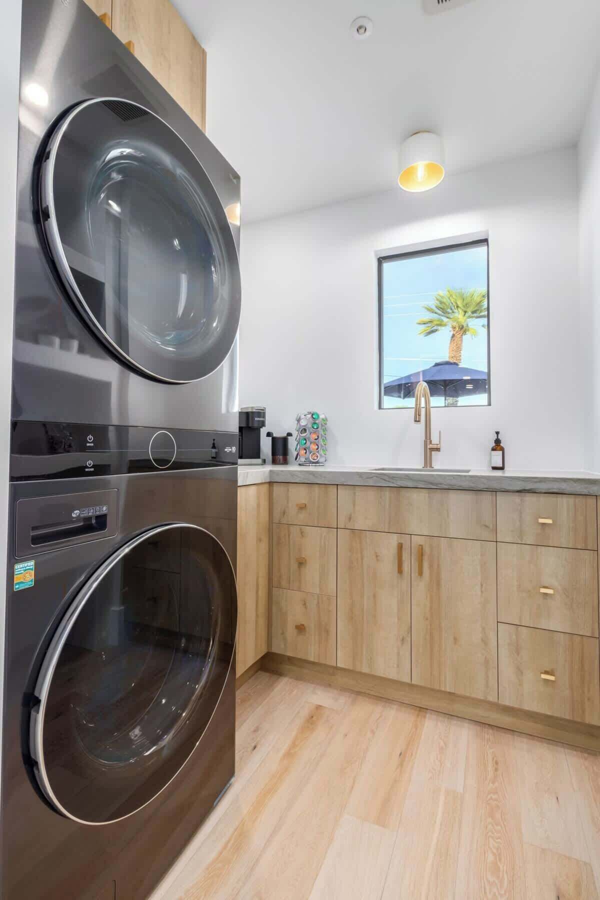 A compact kitchen with a built-in washer and dryer, wooden cabinets, a window with a view of palm trees, and a modern faucet.