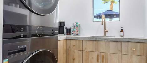 A compact kitchen with a built-in washer and dryer, wooden cabinets, a window with a view of palm trees, and a modern faucet.