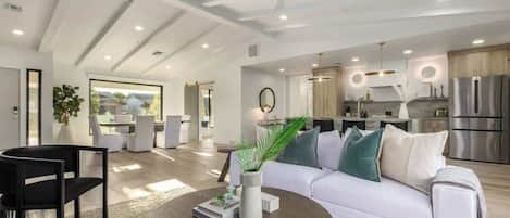 A modern living room with a white sofa, a round coffee table, and a black chair. There is a kitchen area with stainless steel appliances and a dining area with a white table and chairs.