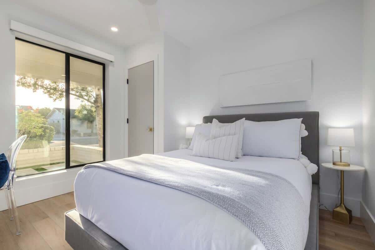 A modern bedroom with a large bed, a sliding glass door leading to a balcony, and a bedside table with a lamp.