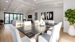 A spacious living area with a large wooden dining table, white upholstered chairs, a large potted plant, and a view through glass doors to the outdoors. - The Kingsley|Custom|Heated Pool|Chef Kitchen|Chess (Scottsdale)