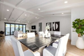 A spacious living area with a large wooden dining table, white upholstered chairs, a large potted plant, and a view through glass doors to the outdoors. - The Kingsley|Custom|Heated Pool|Chef Kitchen|Chess (Scottsdale)