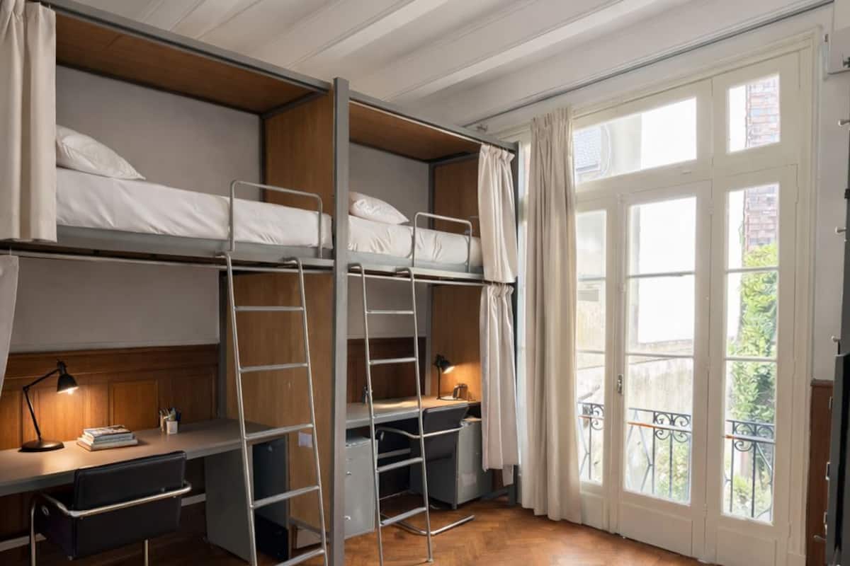 Ein Schlafraum mit Etagenbetten, einem Schreibtisch, einer Leiter und Blick durch ein großes Fenster.