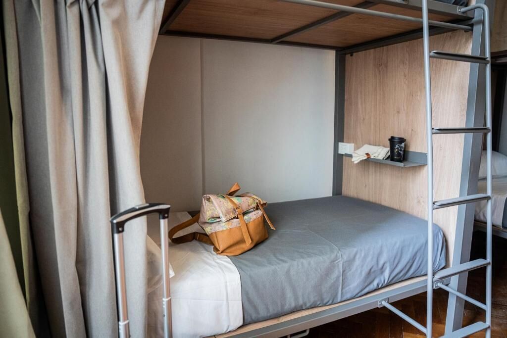 Un lit superposé avec un matelas gris, une échelle en métal, une petite étagère avec une tasse et un sac posé sur le lit.