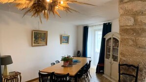 A dining area with a wooden table, black chairs, and a chandelier. There are two framed pictures on the wall and a stone wall on the right.