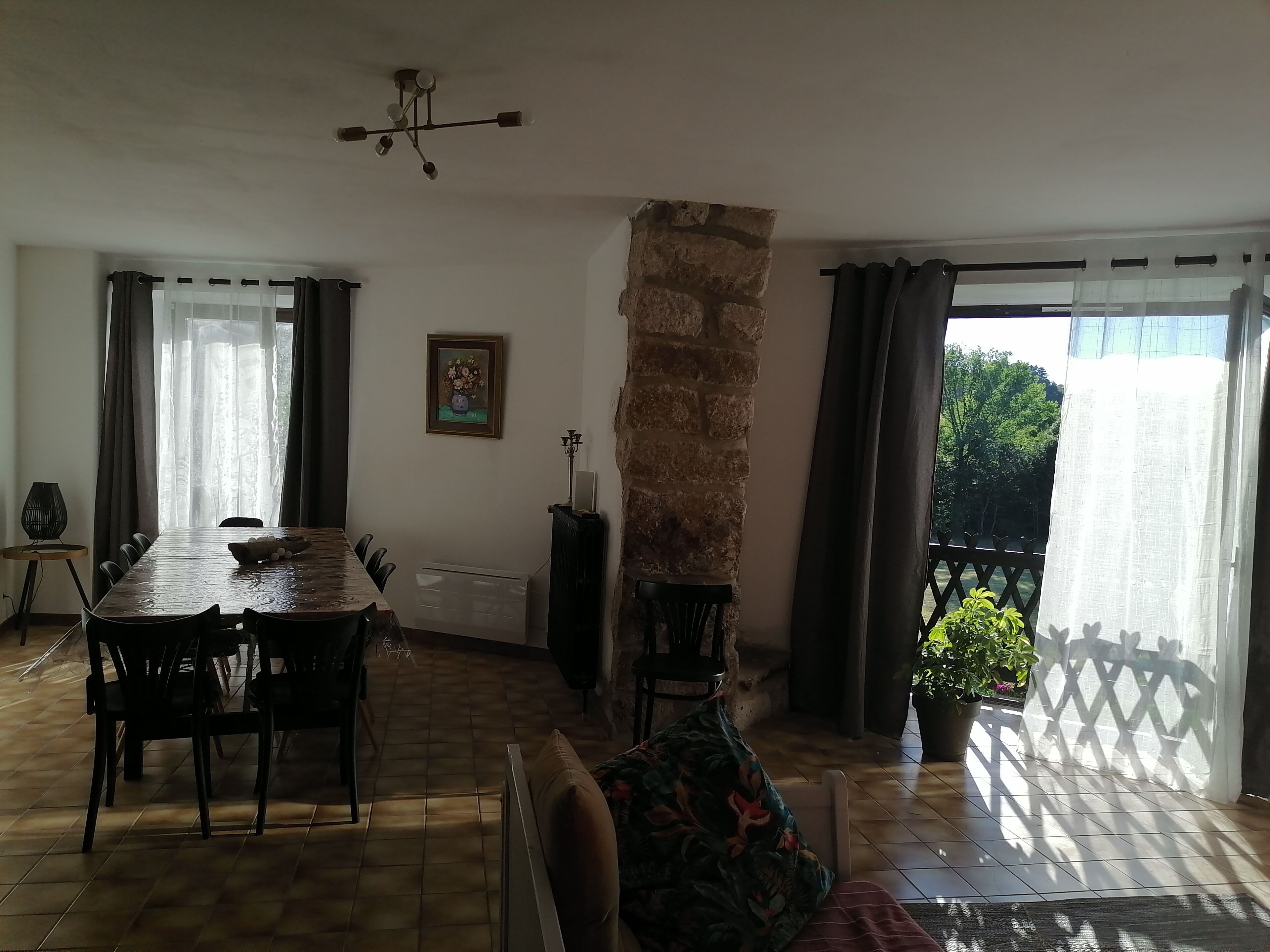 A dining area with a long table and chairs, a stone pillar, and a view through a window with curtains.