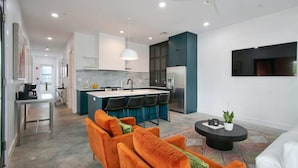 A modern kitchen with a central island, bar stools, and a refrigerator. There is a dining area with orange sofas and a coffee table. - The Garden District's Pied-à-Terre on Prytania | 4 Bedrooms (New Orleans)