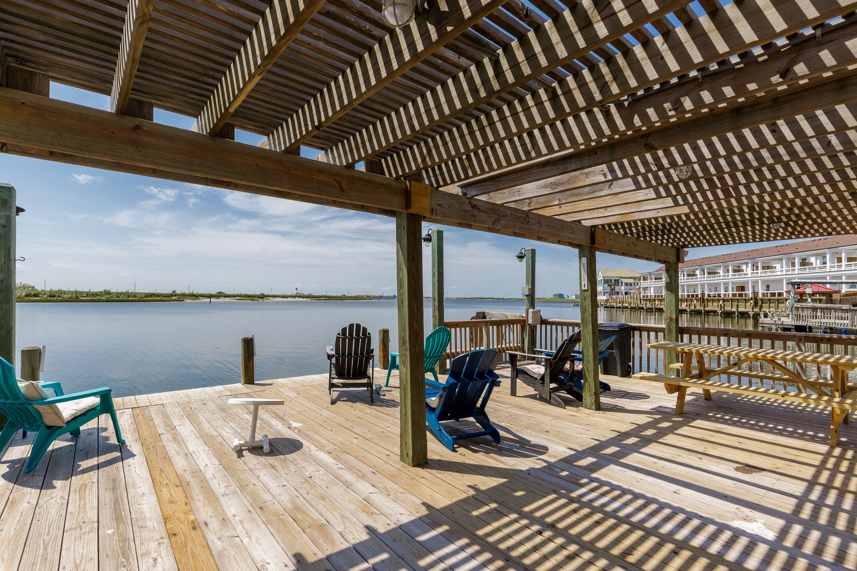 Une terrasse en bois, agrémentée de chaises et de tables, qui offre une vue sur une étendue d’eau calme et un bateau amarré.