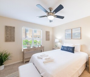 A clean, well-lit bedroom with a bed, bedside tables, a ceiling fan, and a window with blinds.