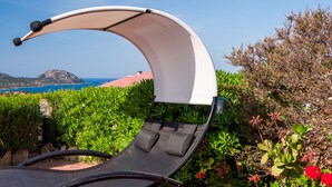 Un transat doté d’un auvent incurvé, placé dans un jardin luxuriant offrant une vue sur la mer et les collines.