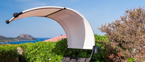 A sun lounger with a curved canopy, positioned in a lush garden with a view of the sea and hills.