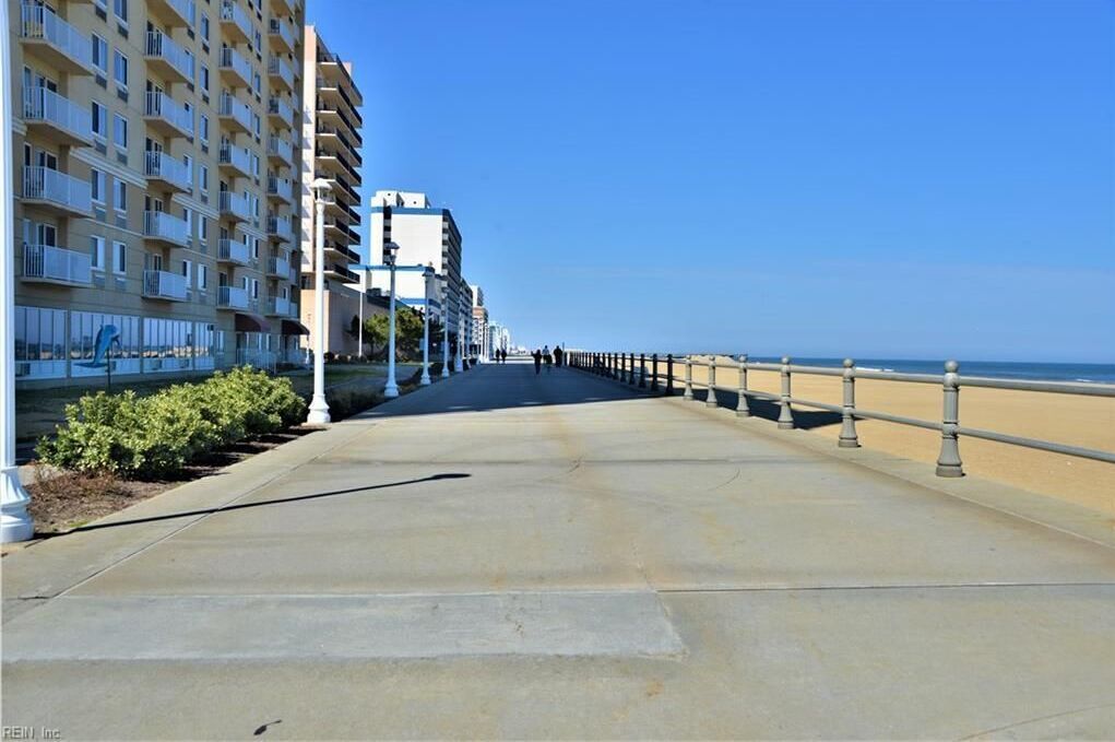 A long, straight pathway leading to a beach, flanked by buildings with balconies and a railing along the water's edge.