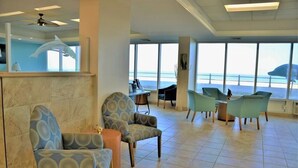 A spacious lobby with a large window overlooking the beach, several seating areas, and a reception desk. - Dolphin Beach condo with indoor pool, ocean views, steps to the beach (Virginia Beach)
