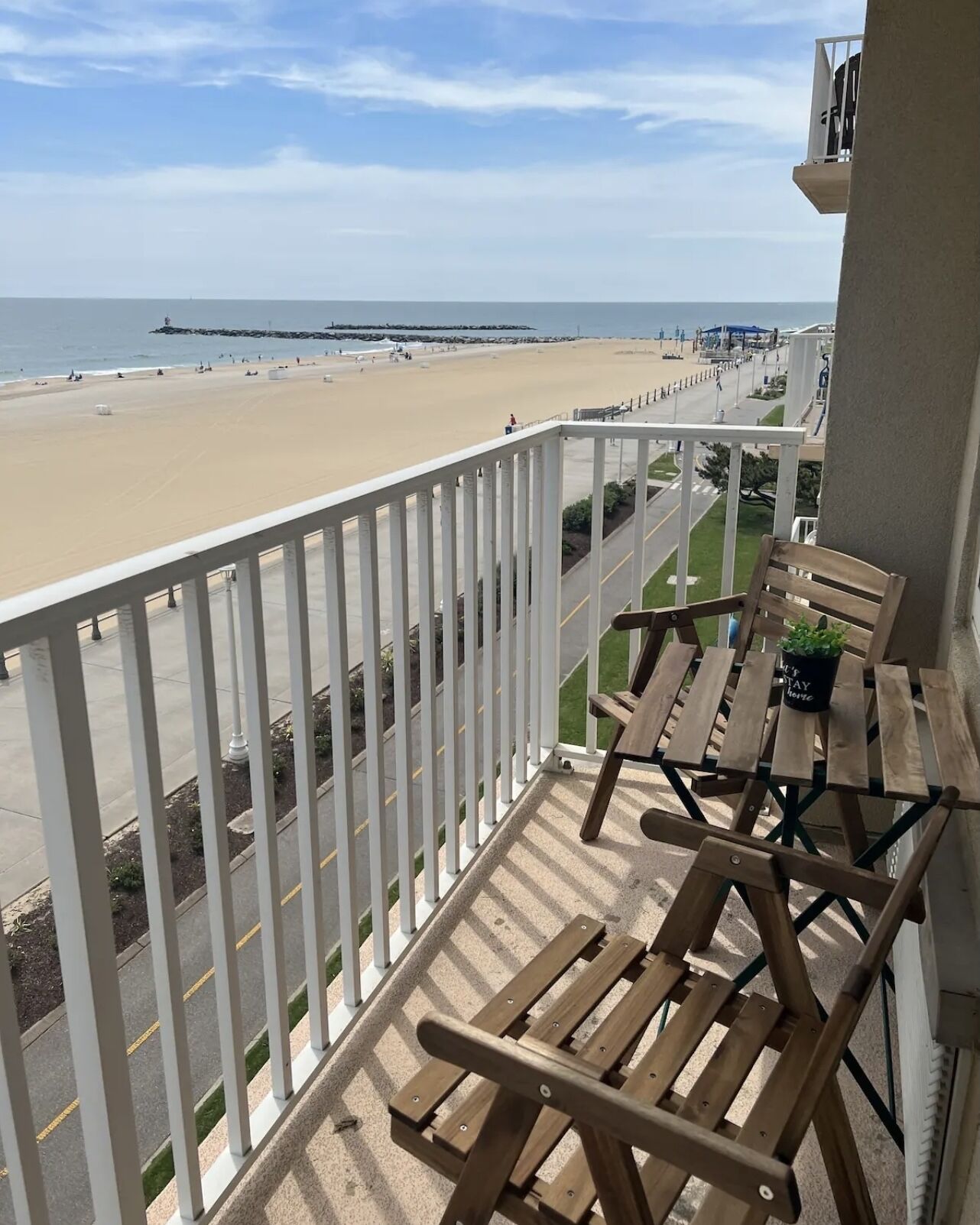 A balcony with wooden chairs and a table, overlooking a beach with a clear view of the ocean and a pier in the distance.