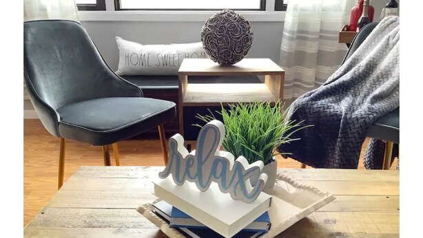Una sala de estar acogedora con una mesa de centro de madera, un cartel decorativo con la palabra 'relax', una planta en maceta y un sillón gris.