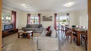 A living room with a sofa, coffee table, TV, and dining area with chairs and a table. - Villa Das Oliveiras, Large grounds, walk to Marina (Albufeira)