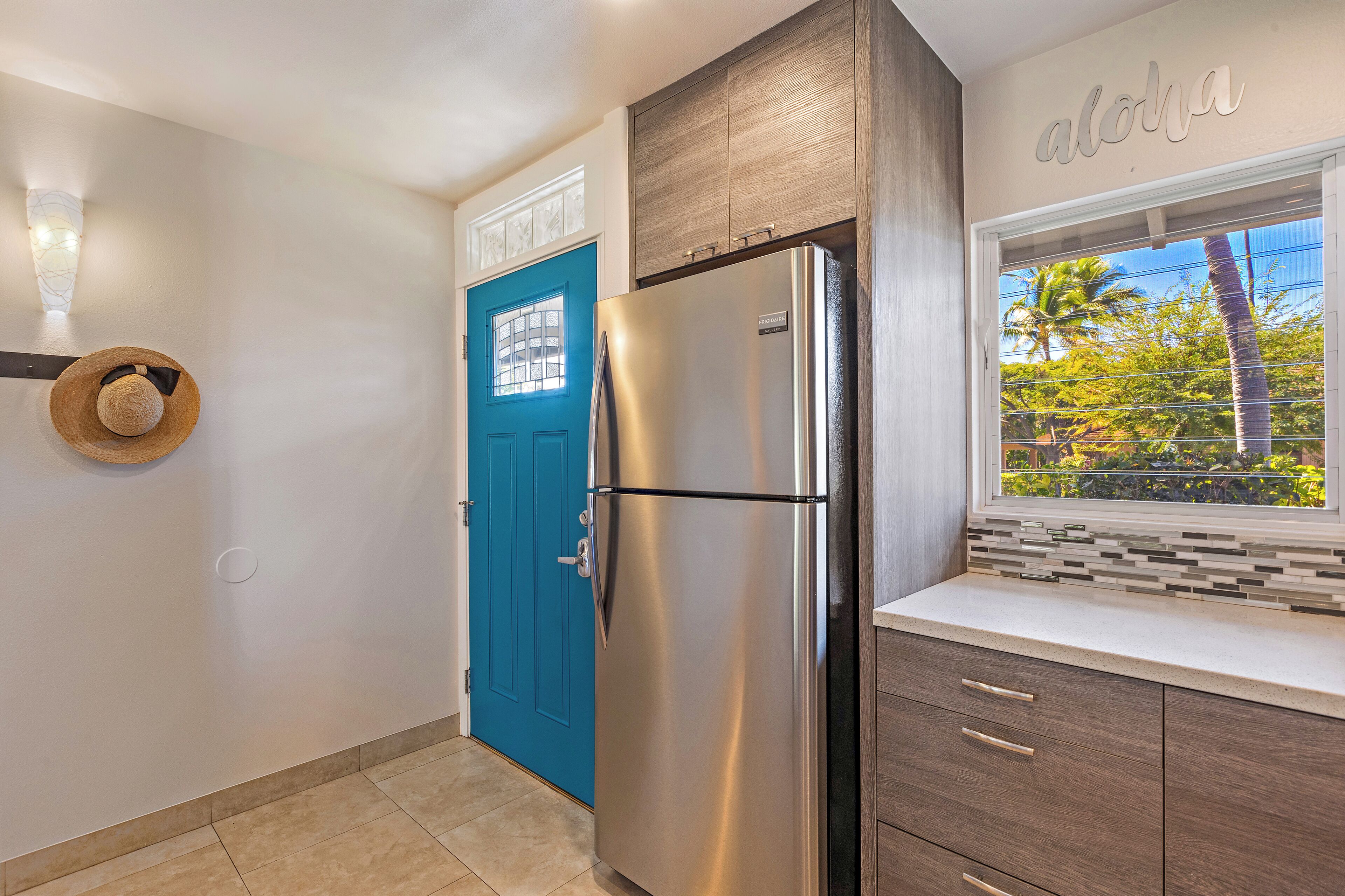 A kitchen with a blue door, a stainless steel refrigerator, and a window with a view of palm trees.