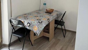 Una zona pranzo accogliente con un tavolo ricoperto da una tovaglia fantasia, due sedie nere, un televisore a parete e una bottiglia d'acqua sul tavolo.