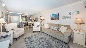 Ein Wohnzimmer mit einem beigefarbenen Sofa, zwei Sesseln, einem Couchtisch und einem gemusterten Teppich. Es gibt einen Essbereich mit Tisch und Stühlen, und eine Küche im Hintergrund.