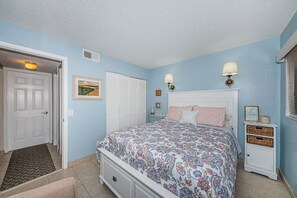 Un dormitorio con cama, mesitas de noche, una mesilla auxiliar, una puerta, una ventana con persianas y una lámpara fijada a la pared.