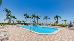 Ein rechteckiger Swimmingpool mit Fliesenbord, umgeben von Liegestühlen und Sonnenschirmen, mit Palmen im Hintergrund.