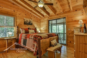 Uma cabana com interior de madeira, uma cama com manta xadrez, um ventilador de teto e uma janela com vista para as árvores.