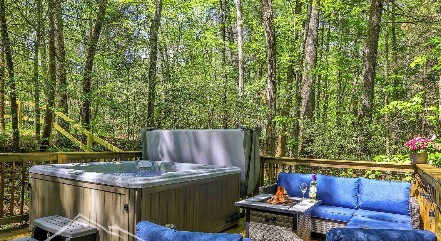 A hot tub on a wooden deck surrounded by wicker furniture and a blue sofa, set in a forested area.