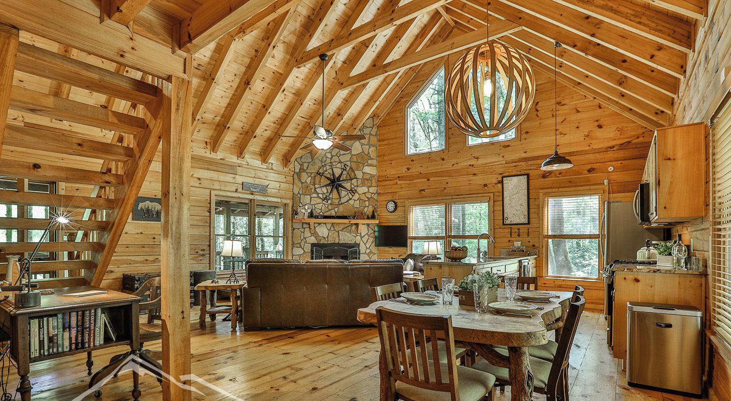 A cabin interior with a stone fireplace, wooden dining table, and a kitchen area.