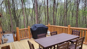 Eine Holzterrasse mit Tisch und Stühlen, umgeben von einem Holzgeländer, eingebettet in einen bewaldeten Bereich.